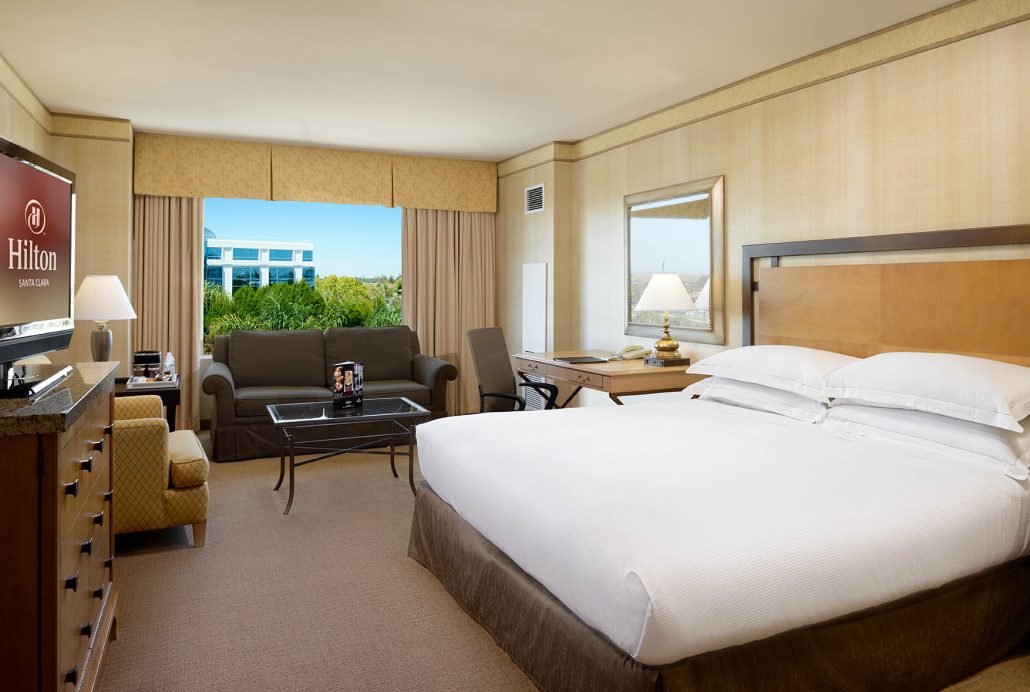 King room with sofabed at Hilton Santa Clara, featuring a spacious bed, seating area, HDTV, workspace, and large window with garden view.
