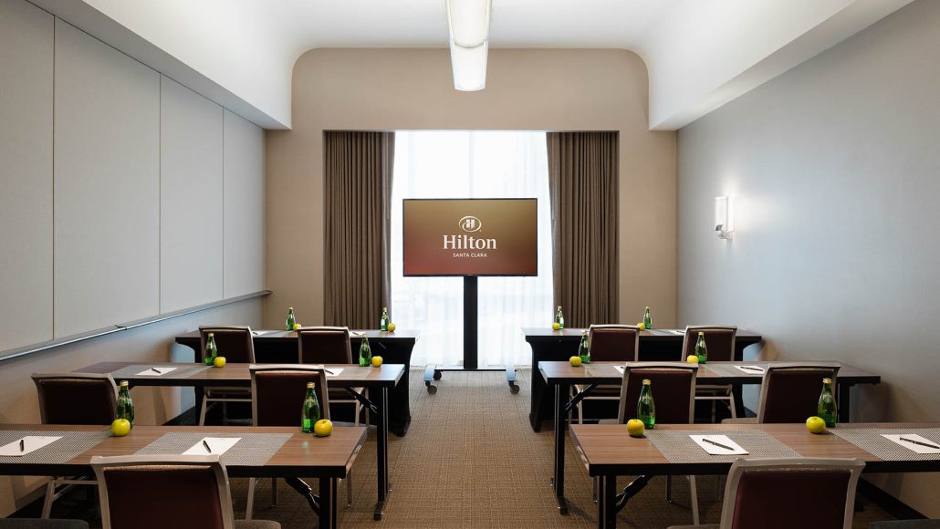Carmel Boardroom at Hilton Santa Clara set up in conference style with tables, chairs, bottled water, and presentation screen; accommodates up to 15 guests